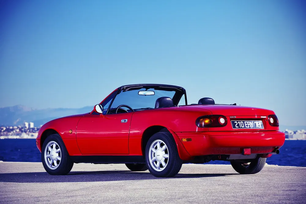 Mazda MX-5 rouge décapotable garée près de la mer avec vue sur la ville.