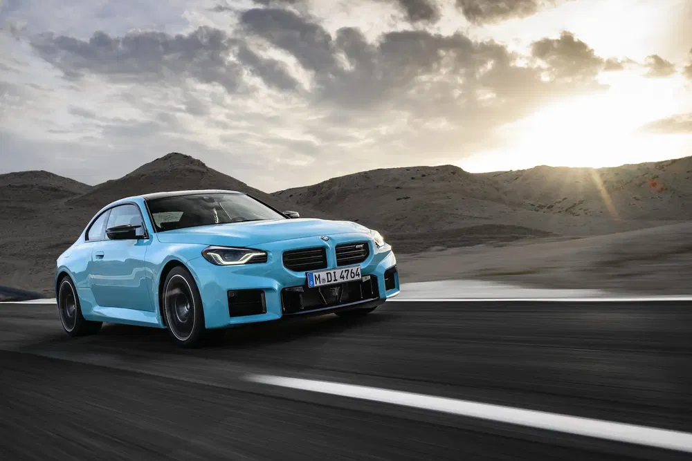 BMW bleu sur route déserte avec montagnes et coucher de soleil en arrière-plan.