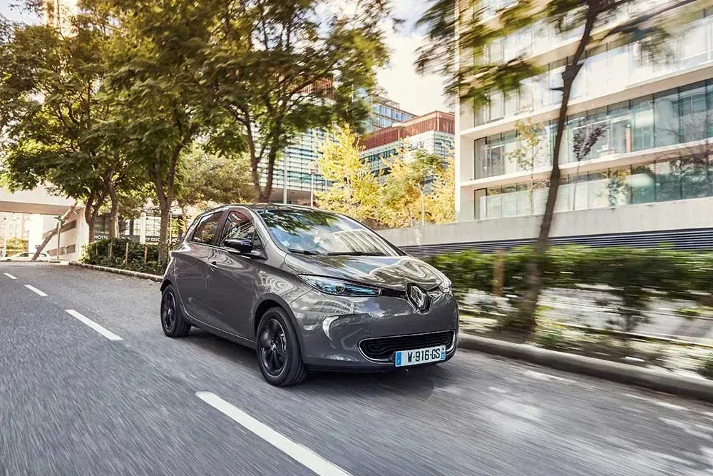 Grijze Renault Zoe rijdt in boomrijke stad met moderne gebouwen op de achtergrond.