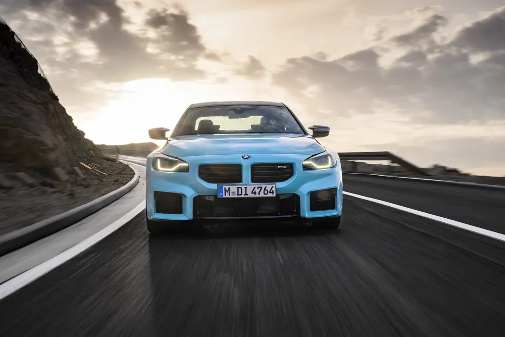 BMW M2 bleu sur route sinueuse au coucher du soleil, vue avant dynamique.