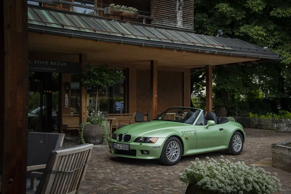 BMW Z3 vert stationnée devant un hôtel en bois, entourée de plantes et mobilier extérieur.