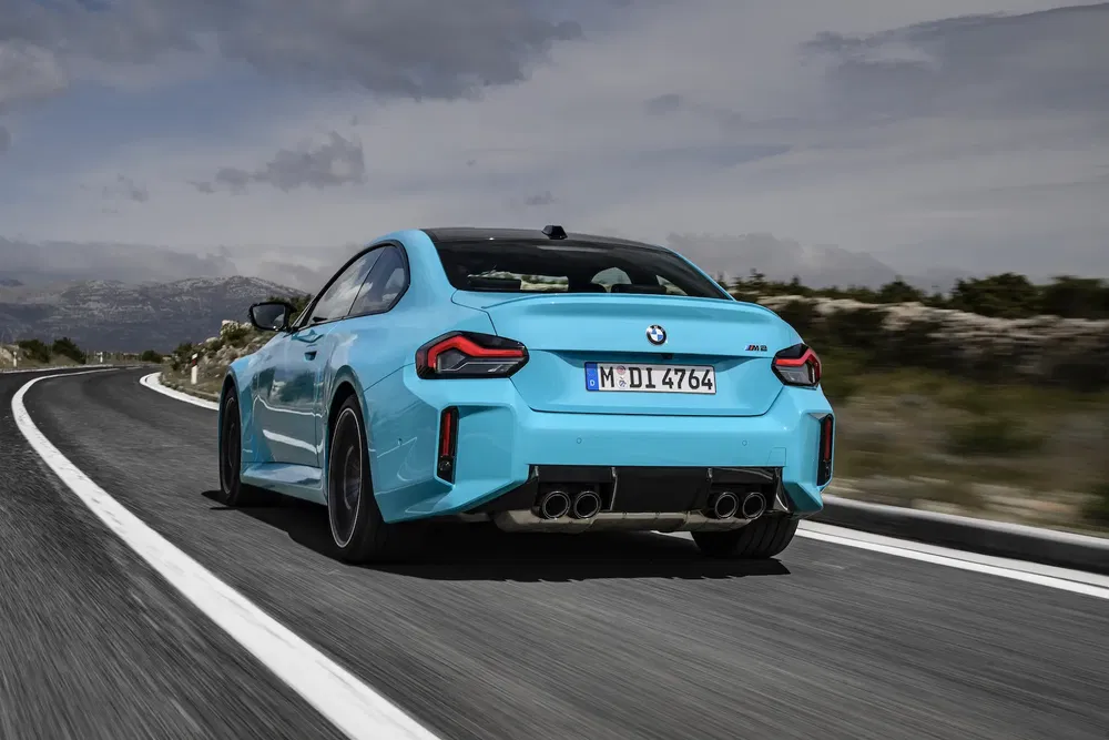 BMW M2 bleue sur route sinueuse, vue arrière dynamique par temps nuageux.