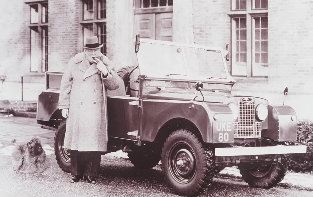 {"fr":"Homme à côté d'une Land Rover Classic, portant un manteau, avec un chien.","nl":"Man naast een klassieke Land Rover, draagt een jas, met een hondje."}