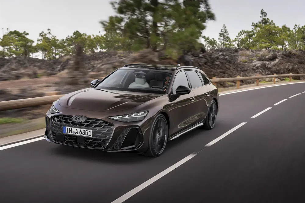 Nouvelle Audi A6 2024 noire élégante roulant sur une route sinueuse bordée d'arbres.