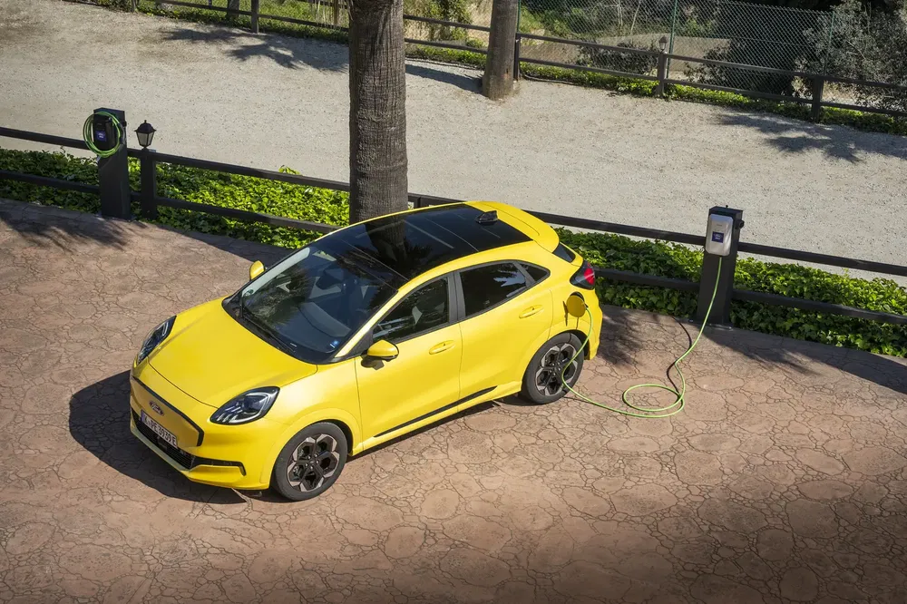 Ford Puma jaune électrique branchée à une station de recharge extérieure.