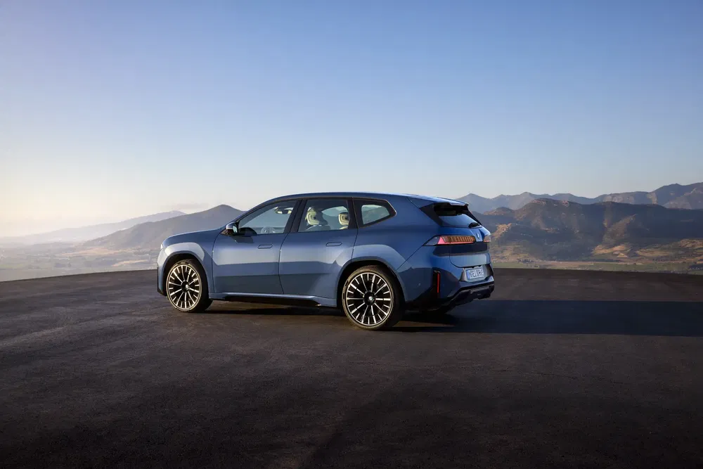 Blauwe BMW iX elektrische auto op panoramische weg, bergen op de achtergrond, heldere lucht.