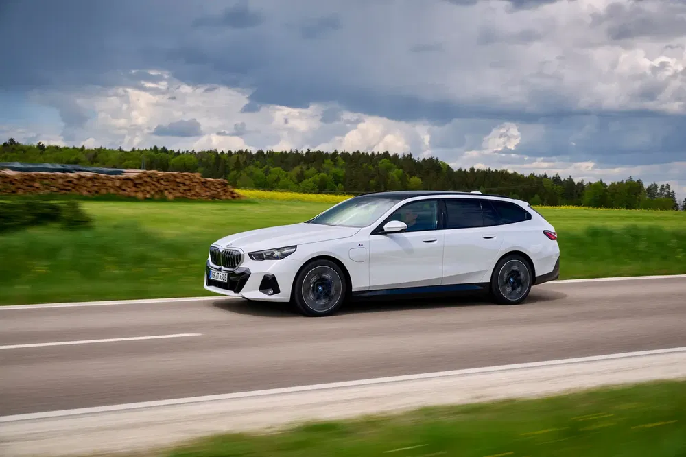 BMW Série 5 Touring blanche roulant sur une route de campagne, ciel nuageux.