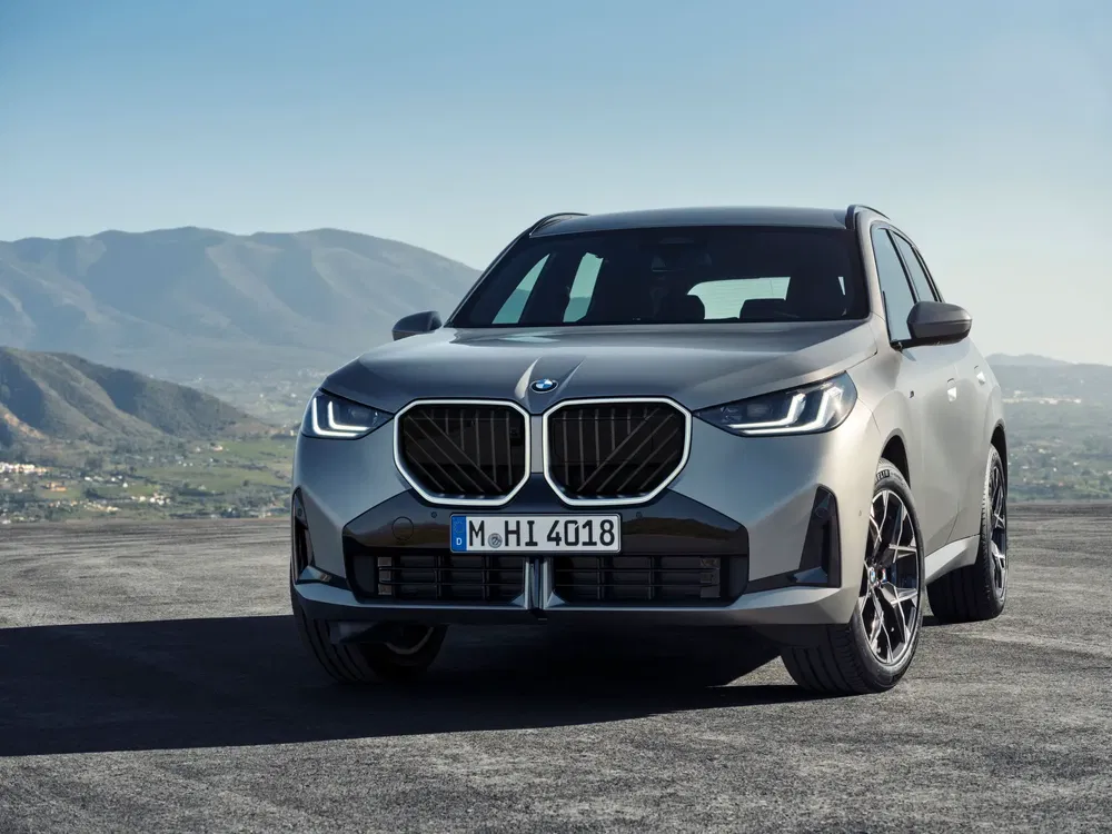 BMW X3 sur une route de montagne, vue avant avec phares allumés.