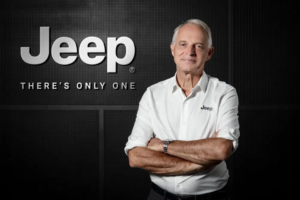 Homme en chemise Jeep blanche debout sous le logo Jeep, fond noir texturé.