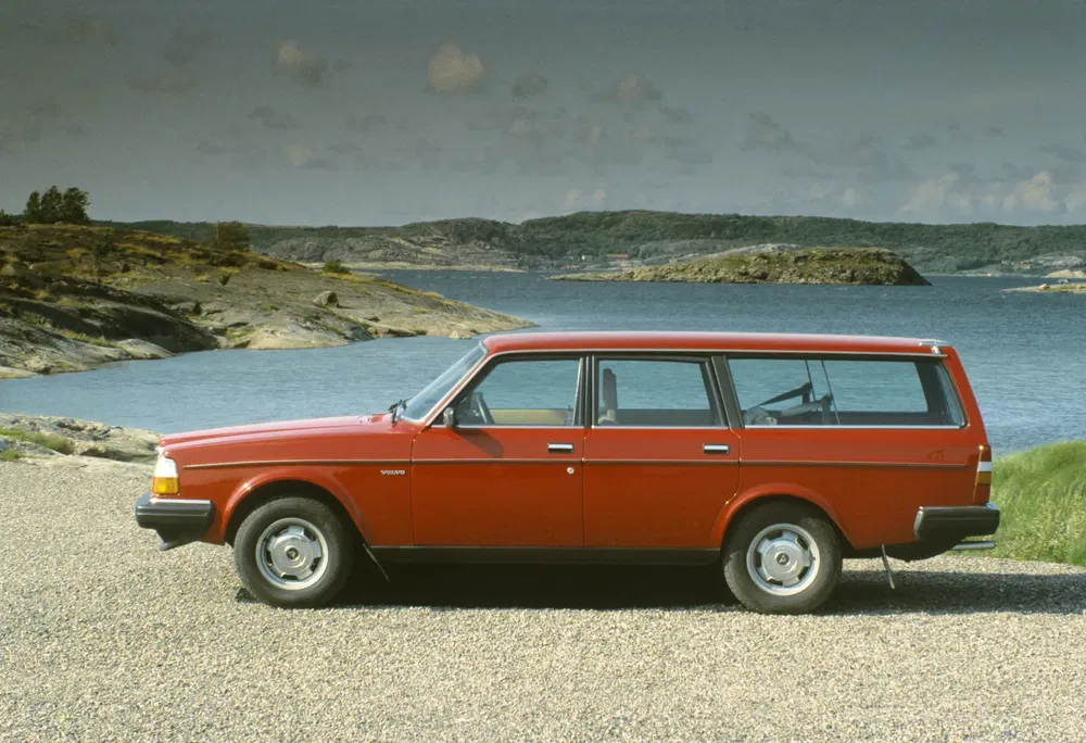 Volvo 245 rouge station wagon, bord de lac avec paysage montagneux en arrière-plan.