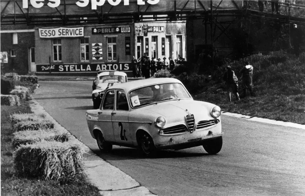 Course automobile avec Alfa Romeo vintage, devant bâtiments publicitaires, années 1960.