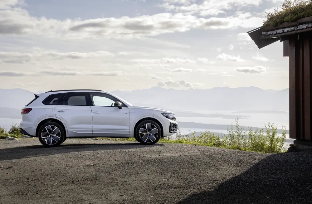 Witte Volkswagen Touareg op een panoramische weg, bergen op de achtergrond onder bewolkte hemel.