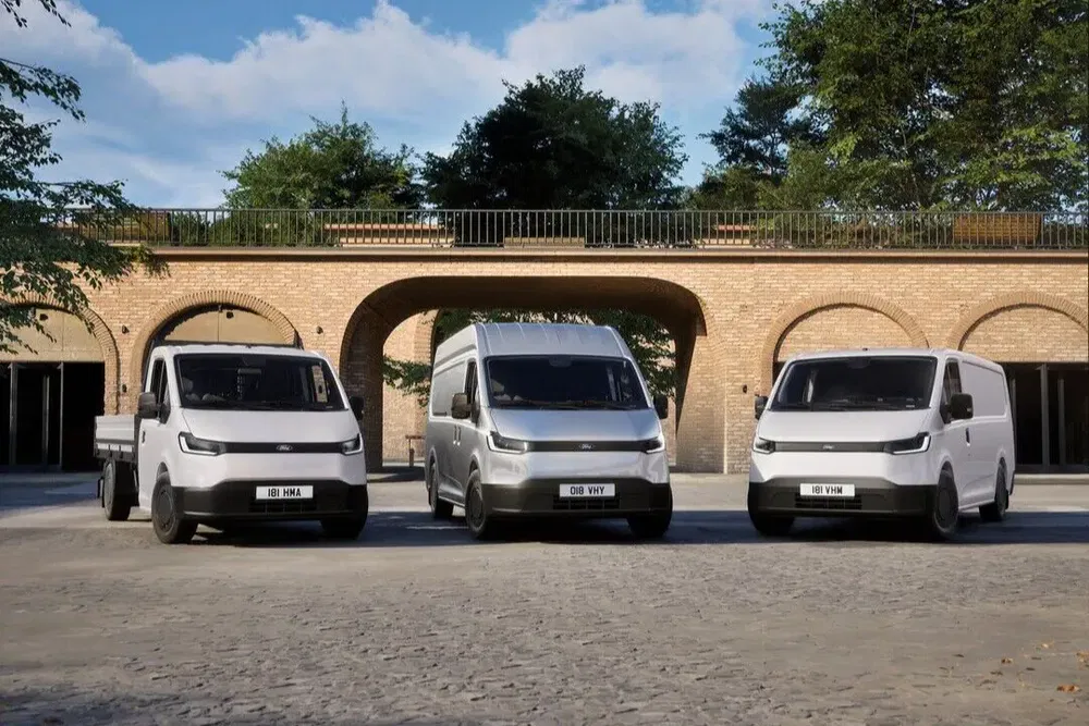 Drie elektrische Ford Transit bestelwagens voor een bakstenen gebouw onder bomen.