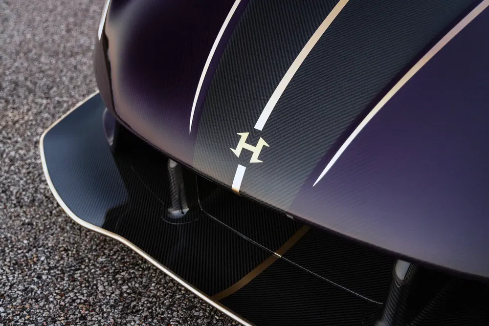 Voorste motorkap hypercar, gouden logo, zwart en paars koolstofverf, sportieve lijnen.