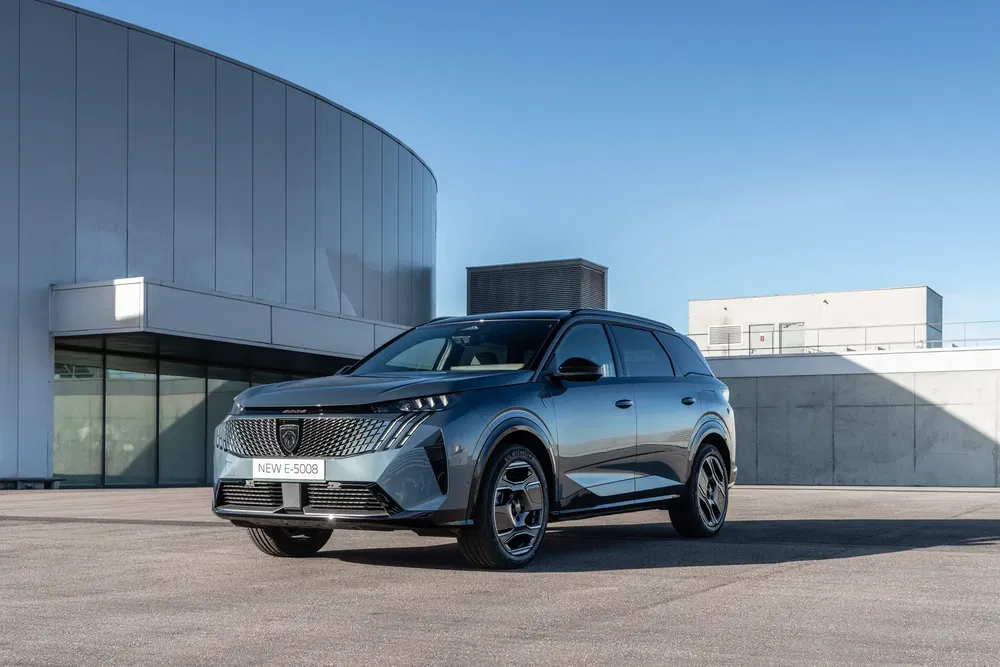 Blauwe Peugeot E-5008 SUV geparkeerd voor een modern gebouw onder een heldere blauwe lucht.