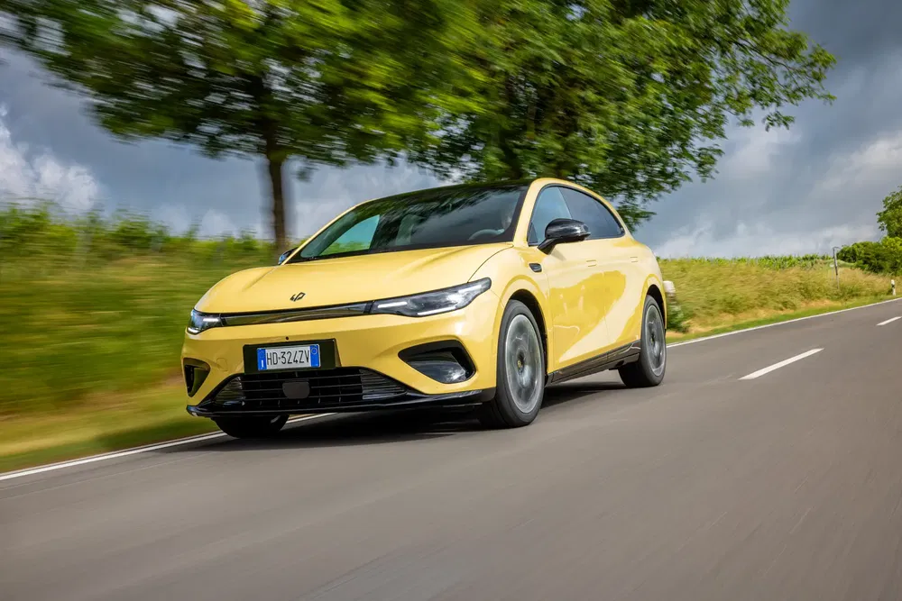 Voiture électrique jaune sur route de campagne, entourée de verdure et ciel nuageux.