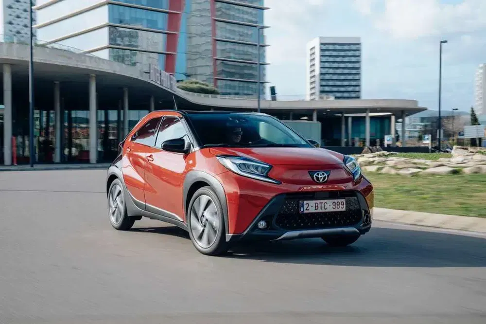 Toyota Aygo X rouge parcourant un quartier urbain moderne, bâtiments en arrière-plan.