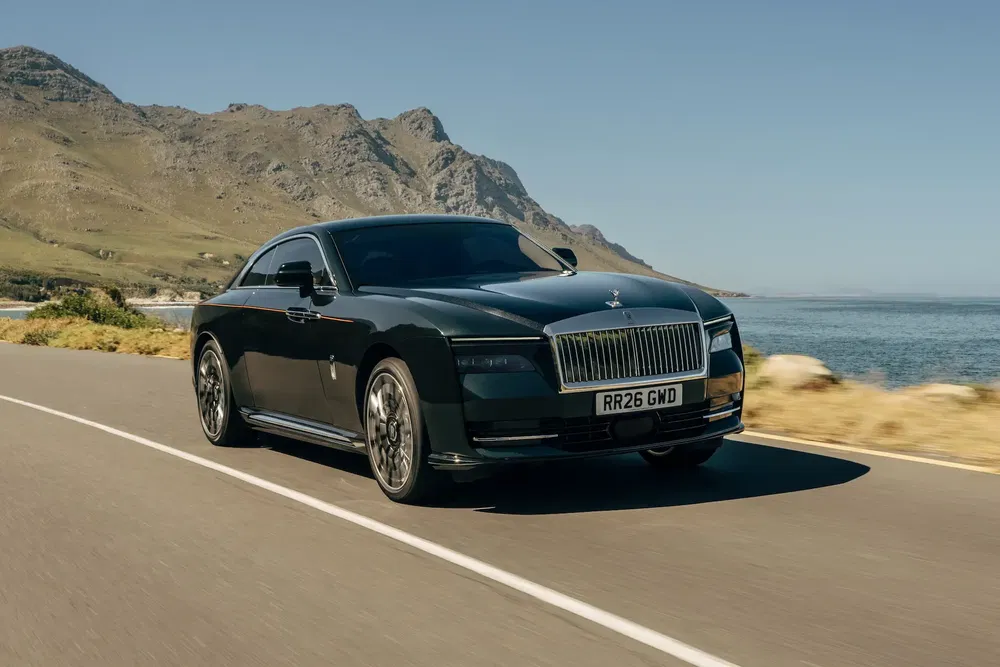 Rolls-Royce coupé noir roulant sur une route côtière, montagne en arrière-plan.