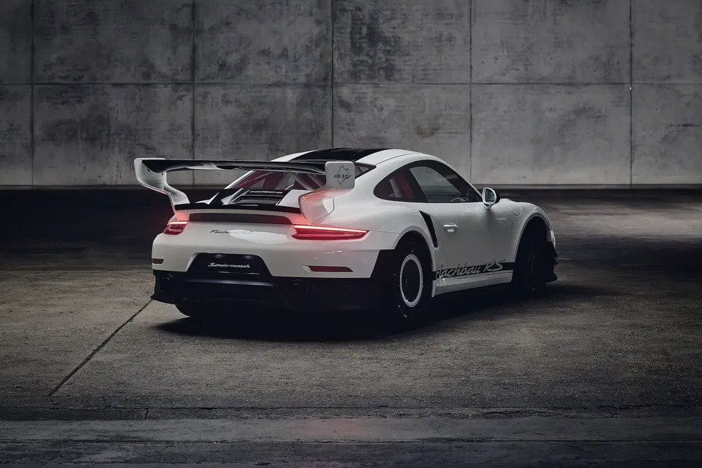 Porsche 911 GT3 RS blanche, vue arrière, aile imposante, stationnée devant un mur brut.