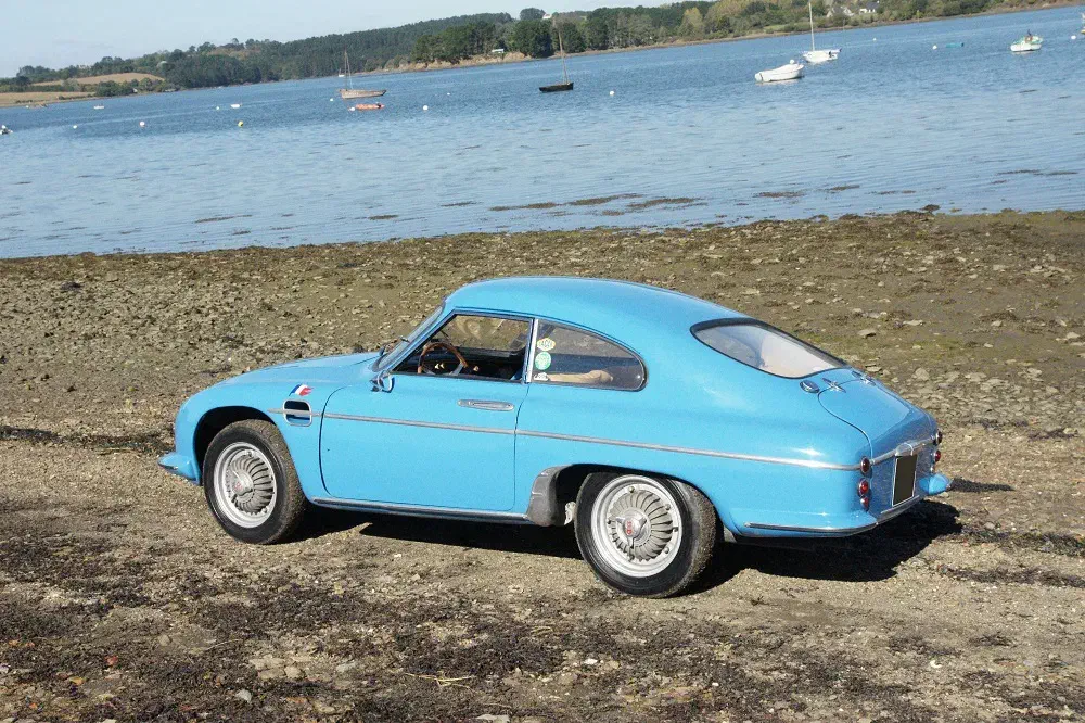Voiture vintage bleue sur la rive, paysage marin avec des bateaux en arrière-plan.