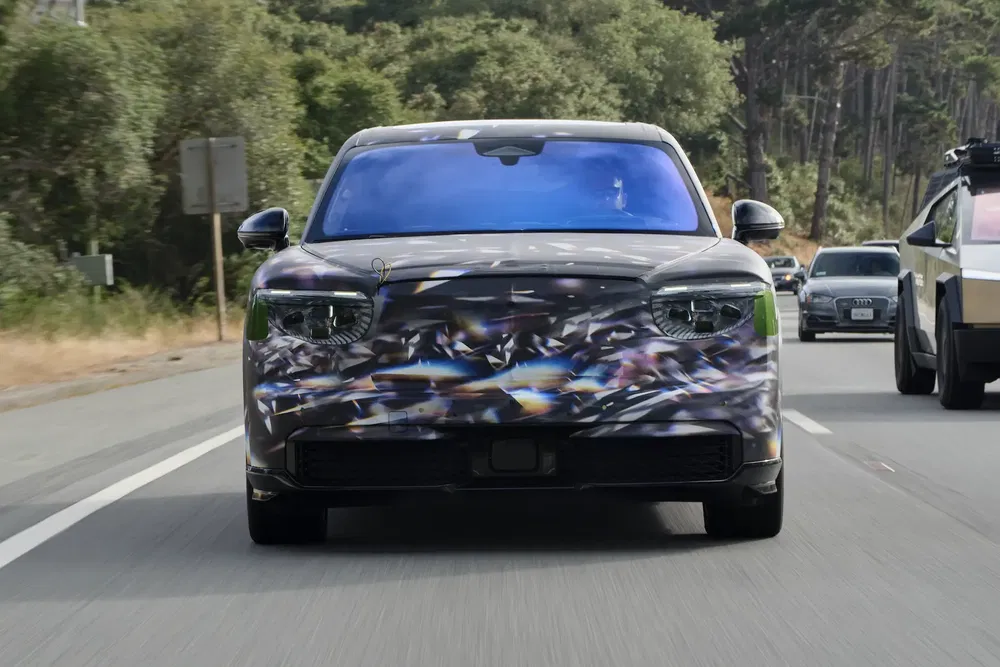 Voiture électrique prototype Kia EV9 camouflée sur route, entourée de paysages verdoyants.