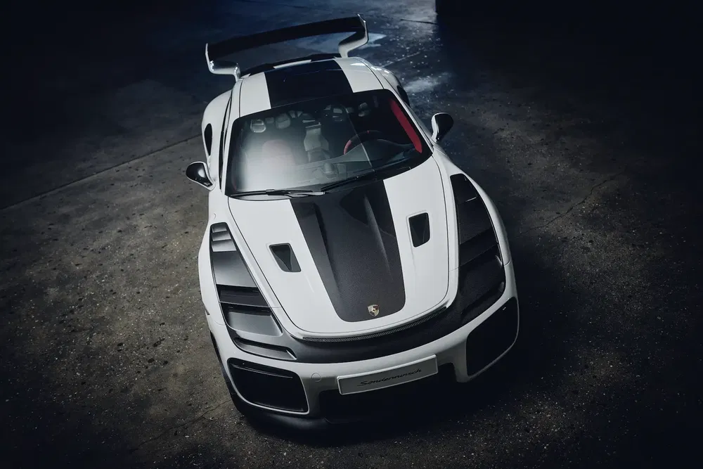 Vue plongeante d'une Porsche blanche avec aileron arrière imposant sur fond sombre.
