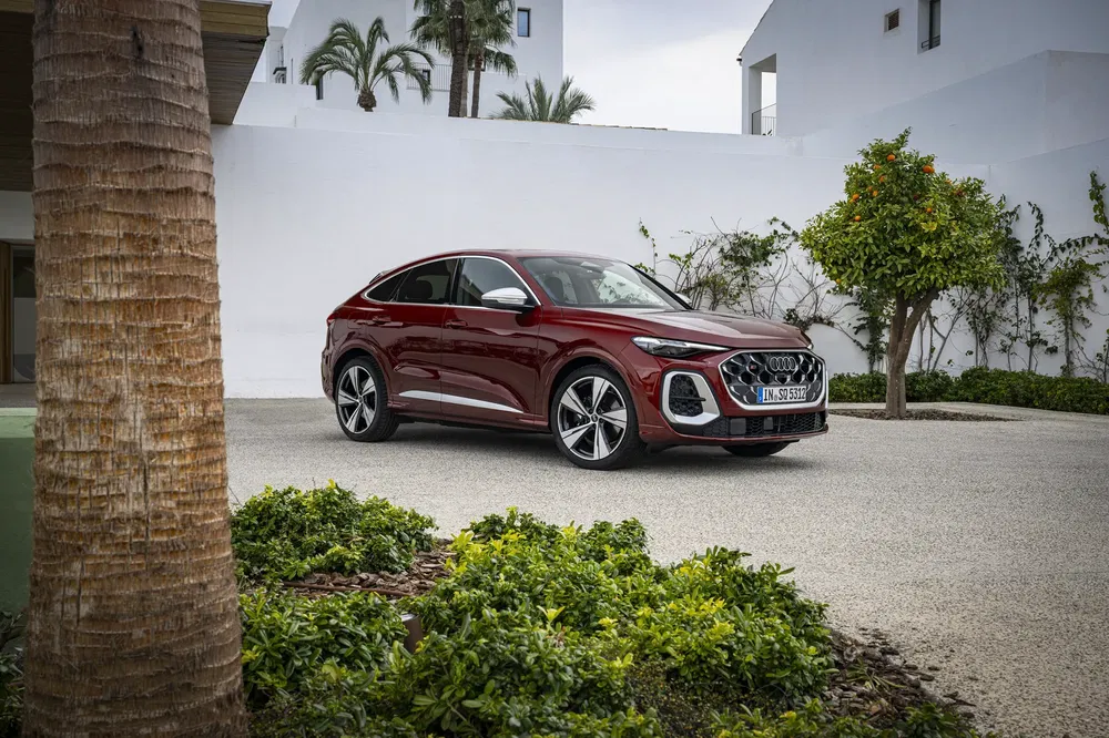Audi Q5 Sportback rouge garée devant un mur blanc avec des plantes et des arbres autour.