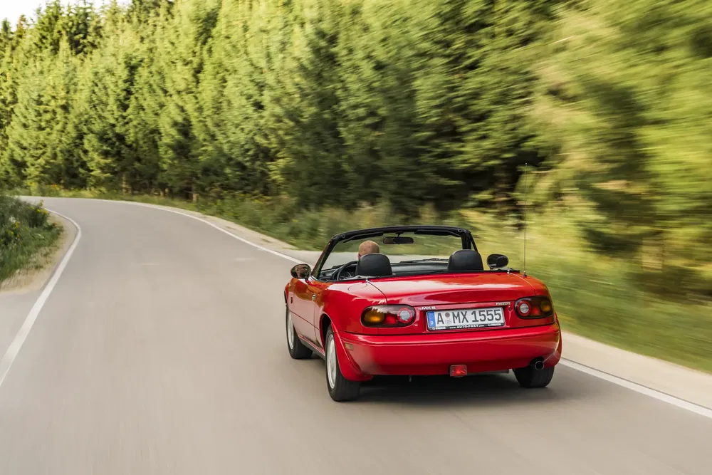 Mazda MX-5 rouge décapotable roulant sur une route bordée d'arbres verdoyants.