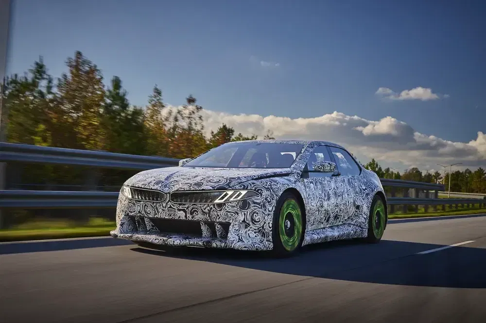BMW prototype auto met camouflage en groene wielen op de weg, bomen op achtergrond.