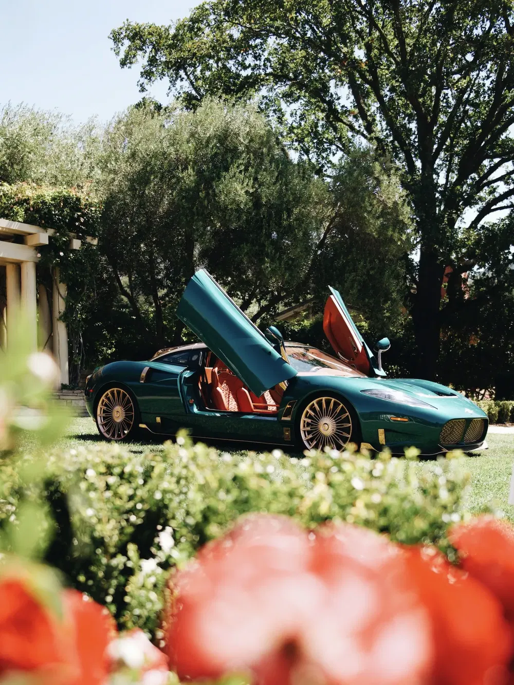 Spyker C8 vert avec portes papillon ouvertes dans un jardin luxuriant.
