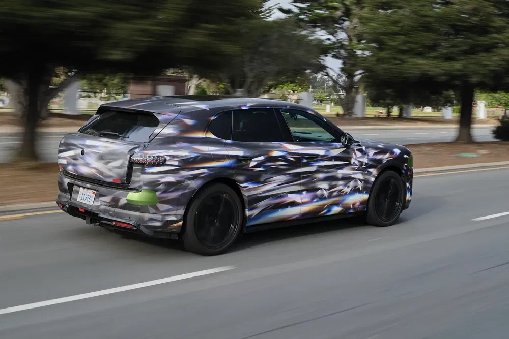 Prototype Porsche Macan électrique camouflé roulant sur une route bordée d'arbres.