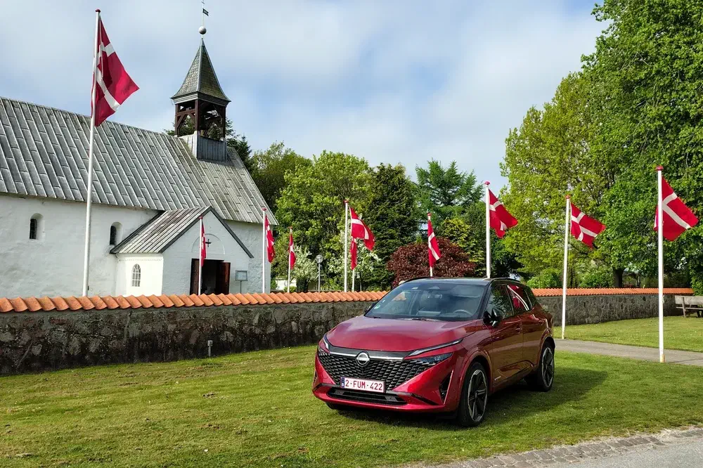 Rode Nissan-auto geparkeerd voor een Deense kerk met omliggende Deense vlaggen.