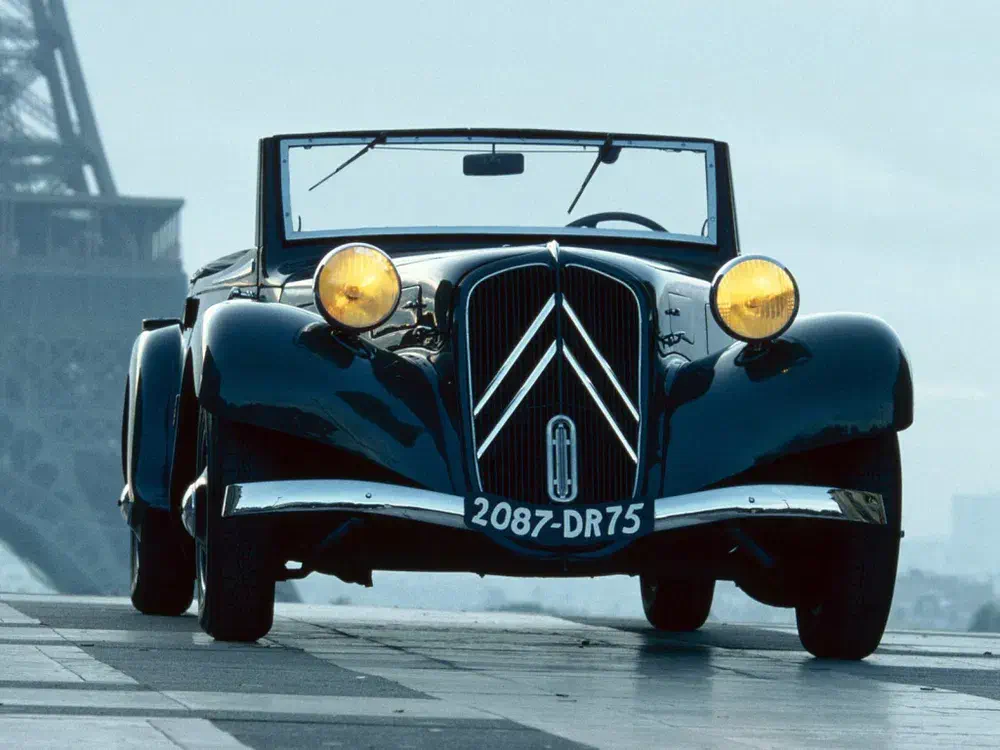 Citroën Traction Avant cabriolet noir devant la Tour Eiffel, phares jaunes allumés.