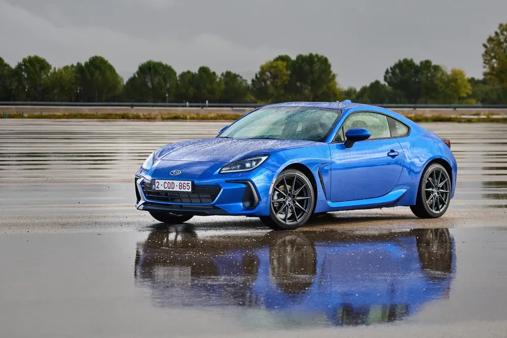 Blauwe Subaru BRZ op nat wegdek, weerspiegelt bewolkte lucht en omringende bomen.