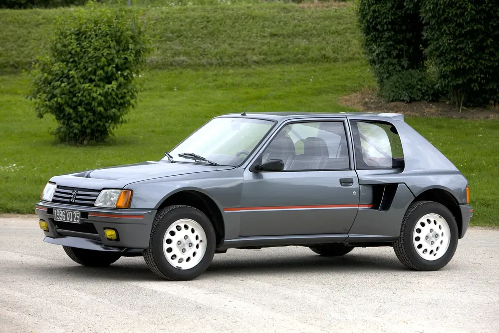 Peugeot 205 Turbo 16 grise des années 80 stationnée sur une route avec arrière-plan verdoyant.