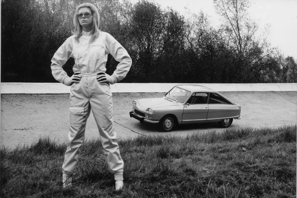 Voiture classique sur piste avec personne en combinaison blanche, Citroën M35 en arrière-plan.