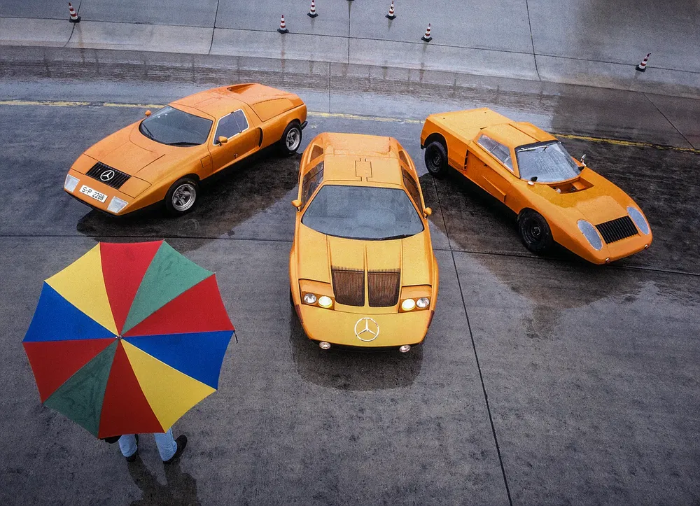 Trois prototypes Mercedes-Benz orange sur une piste humide, avec un parapluie multicolore.