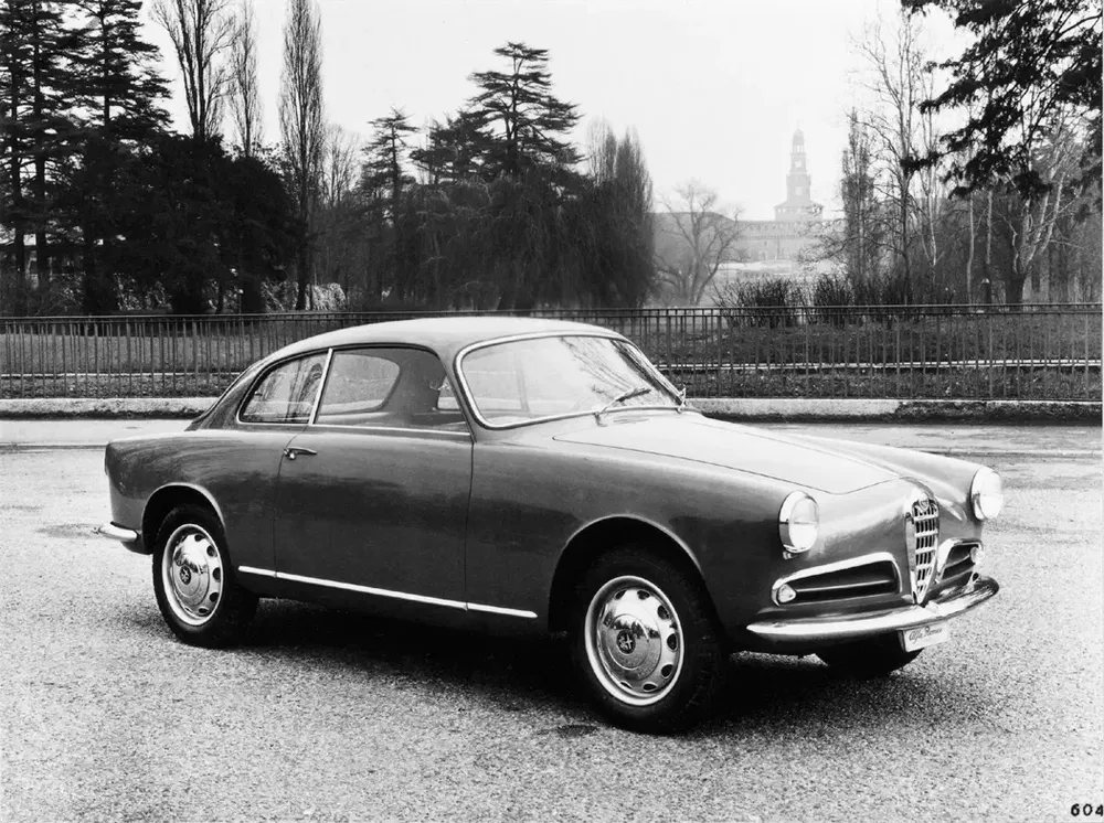 Alfa Romeo classique, probablement la Giulietta Sprint, garée sur une route bordée d'arbres.