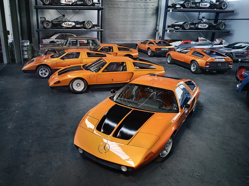 Mercedes-Benz C111 orange dans un garage avec prototypes historiques alignés, ambiance rétro.