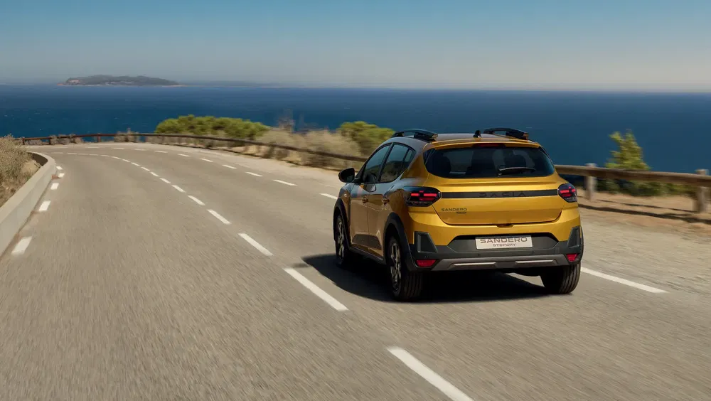 Gele Dacia Sandero Stepway rijdt over kustweg met uitzicht op de oceaan.