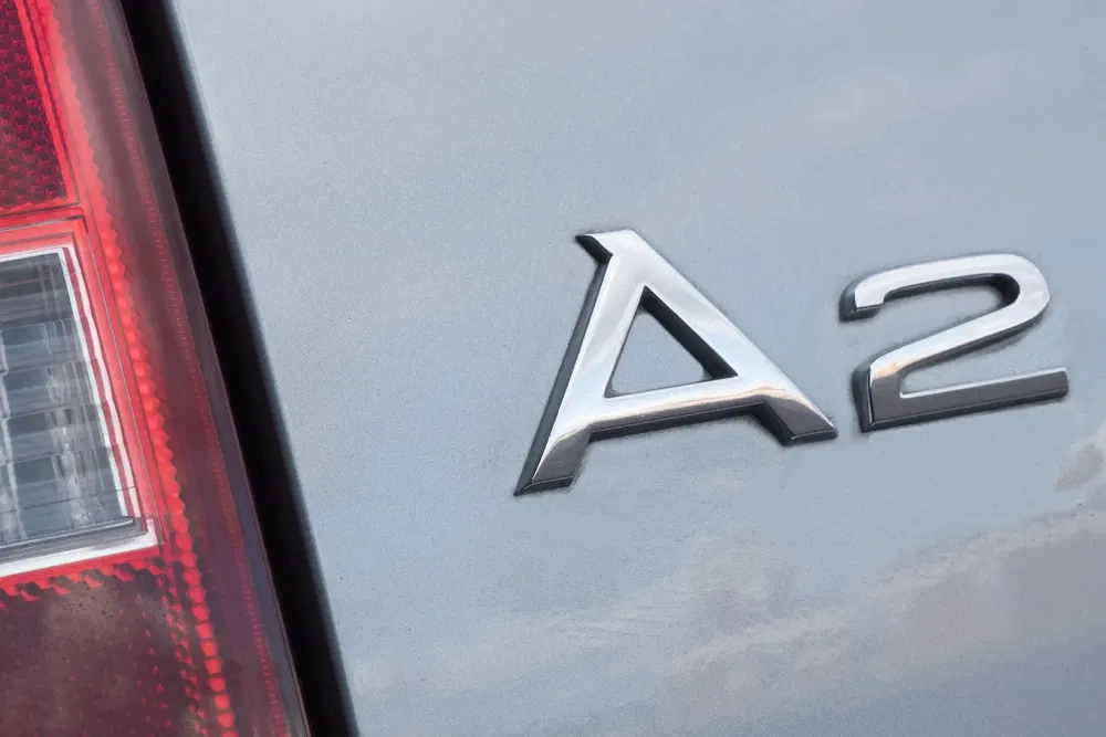 Logo Audi A2 argenté sur carrosserie grise avec phare arrière en arrière-plan.