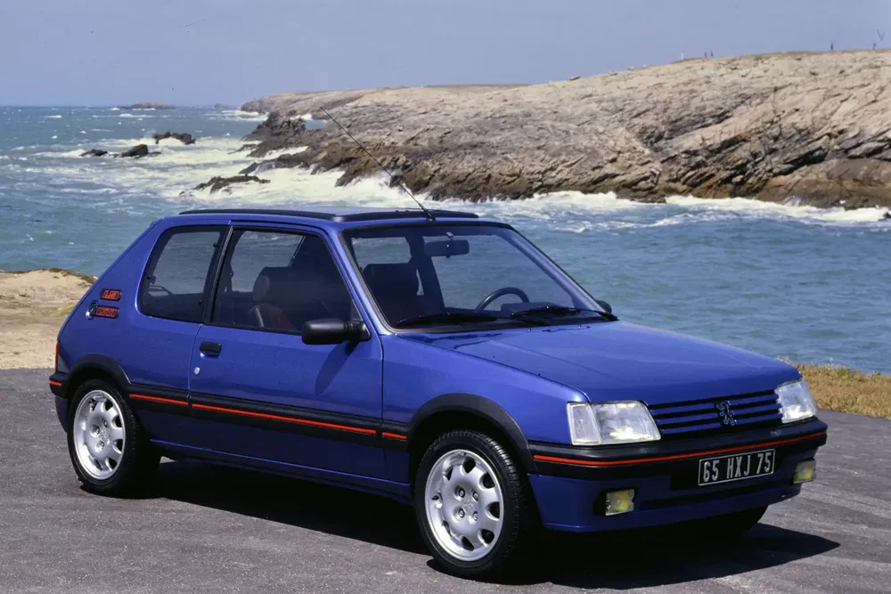 Peugeot 205 GTI bleue au bord de la mer, paysage rocheux et océan agité.