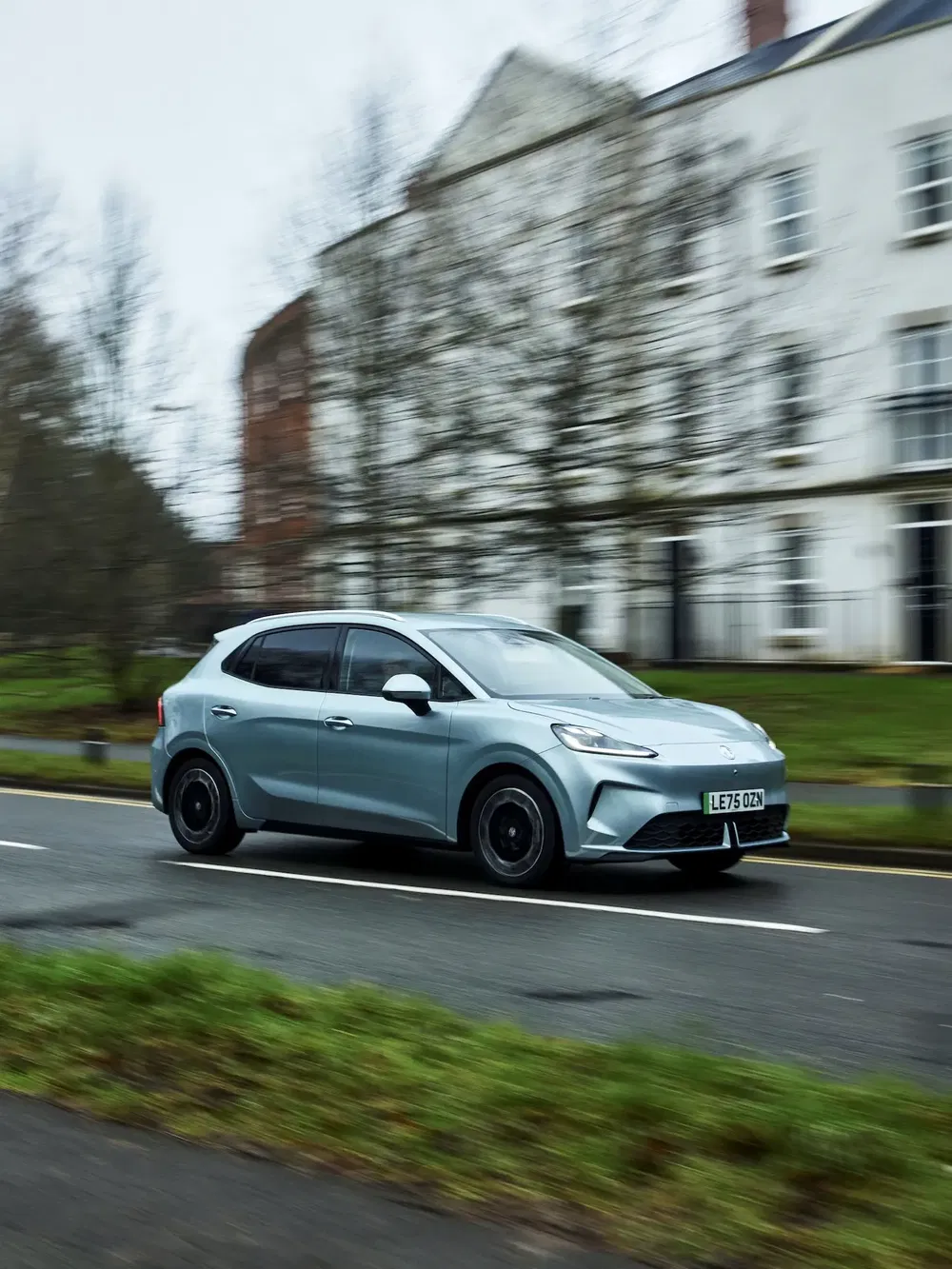 Zilveren elektrische MG4 auto rijdt op natte stadsweg, met wazige gebouwen op de achtergrond.