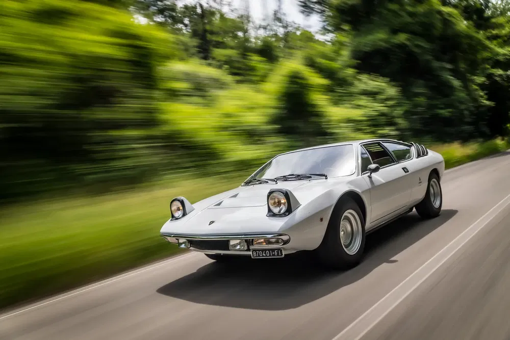 Lamborghini Urraco argentée en mouvement rapide sur une route bordée de verdure.