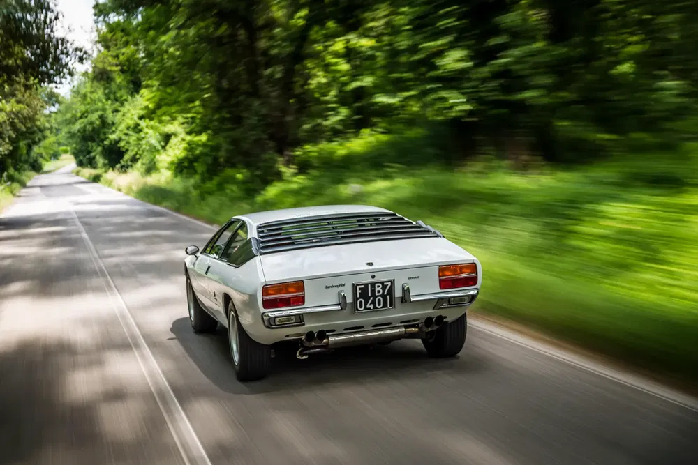 Lamborghini Jarama blanc roulant sur route boisée en été.