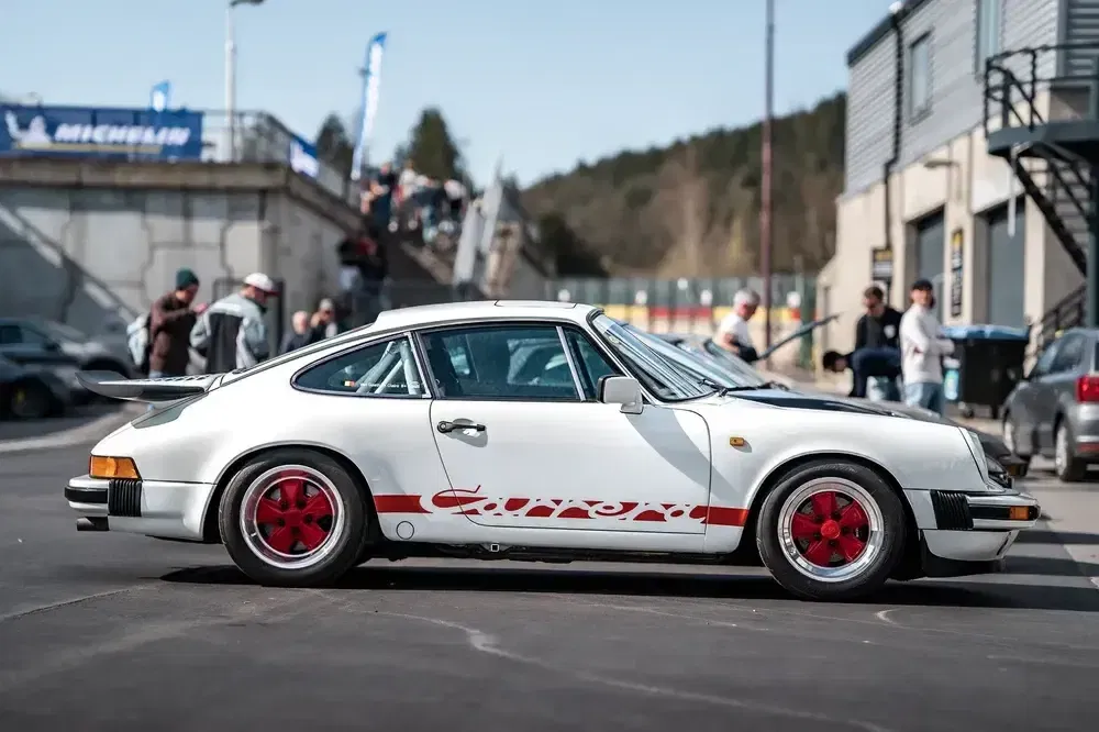 Porsche 911 Carrera classique blanche avec roues rouges, stationnée près d'un circuit.