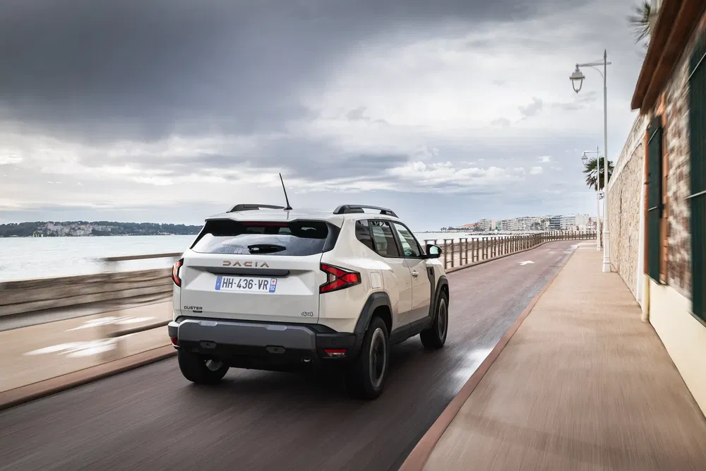 Dacia SUV Duster blanc roulant sur une route côtière, ciel nuageux en arrière-plan.
