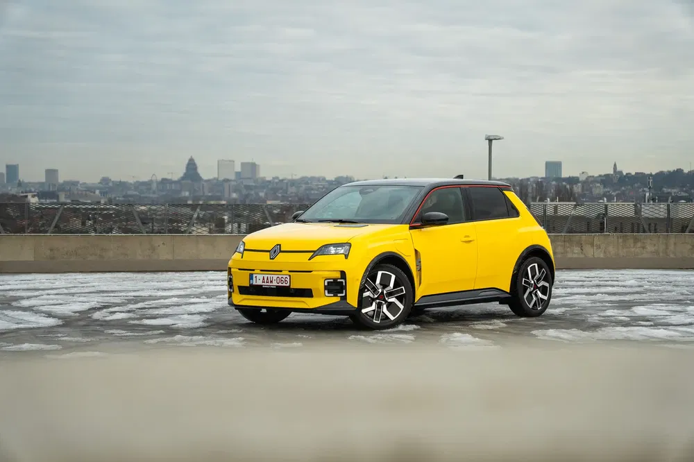 Renault 5 électrique jaune sur un parking urbain avec panorama de ville en arrière-plan.