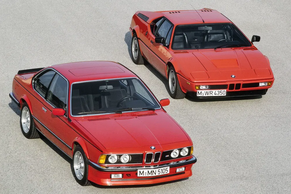 Deux voitures BMW rouges classiques, modèle M1 et Série 6, stationnées sur du béton.
