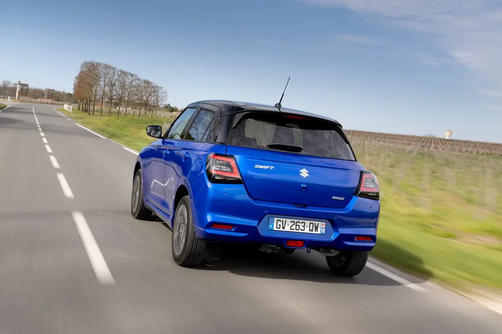 Blauwe Suzuki Swift rijdt op zonnige landelijke weg.